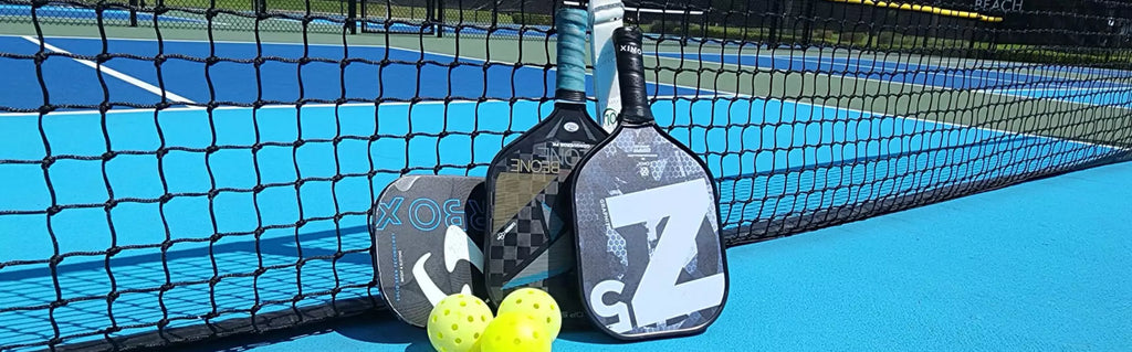 iam-pickleball.com:  A collection of pickleball paddles leaning on a pickleball net.   All selection of pickleball rackets and paddles.