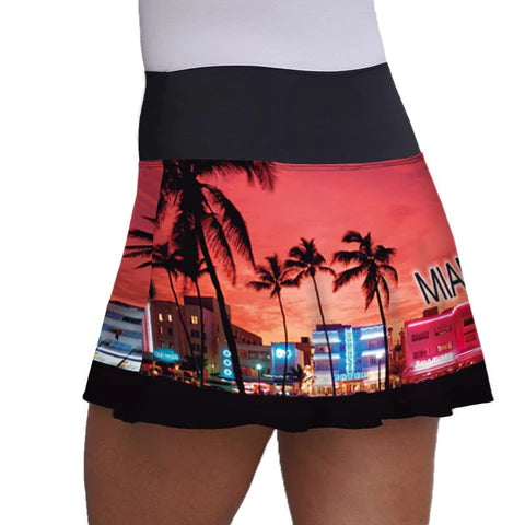 SPORT: PICKLEBALL. Shop Lacoa Sports clothing at "iamracketsports", Miami, FL, USA.  Side on view of Miami Beach Skirt with wrap around print on black of sunset on Miami boulevard with vibrant red,  orange and yellow sky, palm trees in silhouette and lit up buildings.