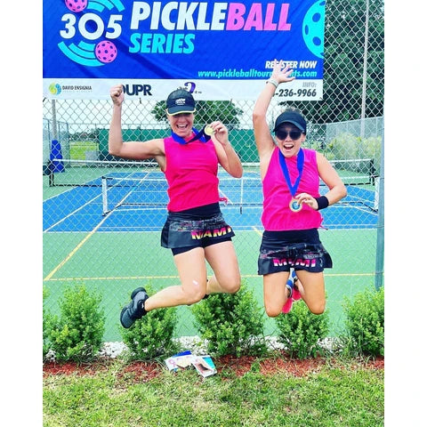 SPORT: PICKLEBALL. Shop Lacoa Sports clothing at USA's premier Racket and Paddle Sports store, "iamracketsports". Two female pickleball players jumping holding medals against court background wearing Lacoa's black MIAMI SKYLINE Skirt printed with Miami Skylines  described in text and the city name shown, using vibrant neon yellow, pink and white.
